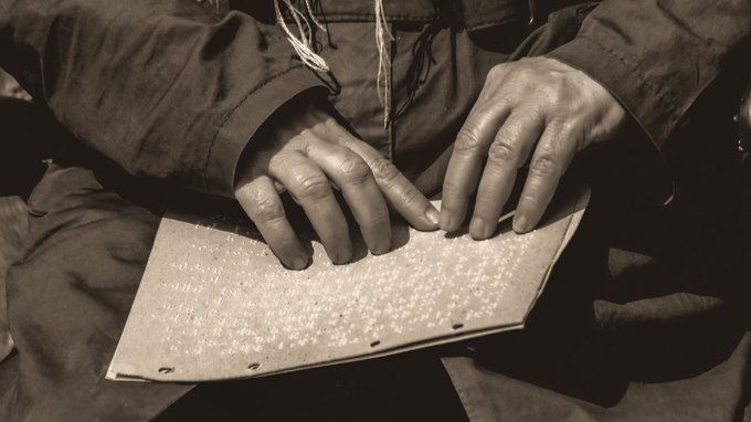 A close-up sepia-toned photograph of a blind person reading braille, with their hands resting on a braille page over their lap.