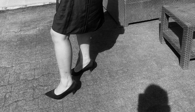 A black and white photograph showing a person's legs in dark high heels and a skirt walking on pavement, with a wicker table visible to the right and the photographer's shadow cast in the foreground.