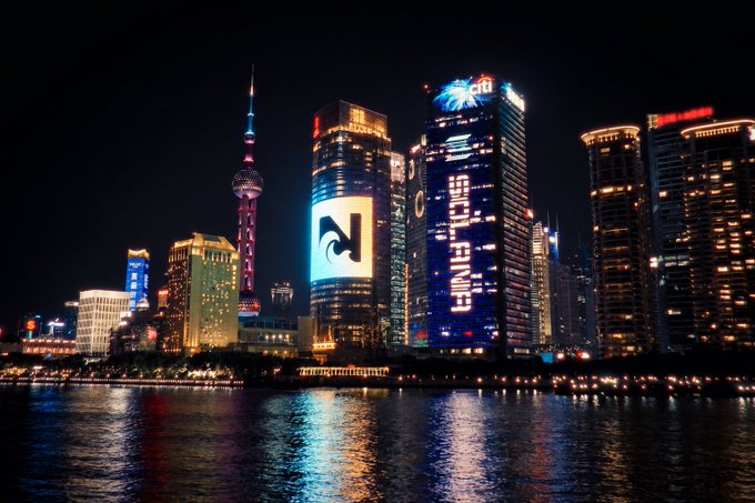 A nighttime photograph of the Shanghai skyline, prominently featuring the Oriental Pearl Tower and illuminated skyscrapers reflecting on the Huangpu River.