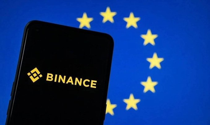 The image shows a smartphone displaying the Binance logo (in yellow text) against a blurred background of the European Union flag's circle of yellow stars on a blue field. This visual pairs with the post about Binance suspending its services in several EU countries starting July 1, 2026, due to incomplete MiCA licensing requirements.