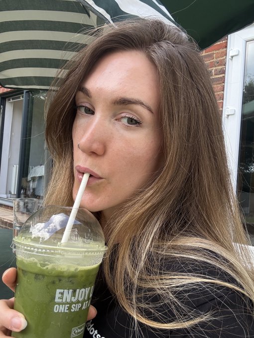 A young woman with long brown hair sipping a green iced drink through a straw from a clear plastic cup labeled 'ENJOY ONE SIP AT A...' while sitting under a green and white striped awning.