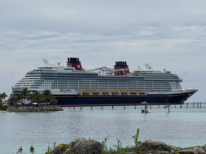 A large Disney Cruise Line ship is docked at a tropical port, featuring the classic black hull with red funnels bearing the Mickey Mouse logo, and yellow lifeboats lined along its side.