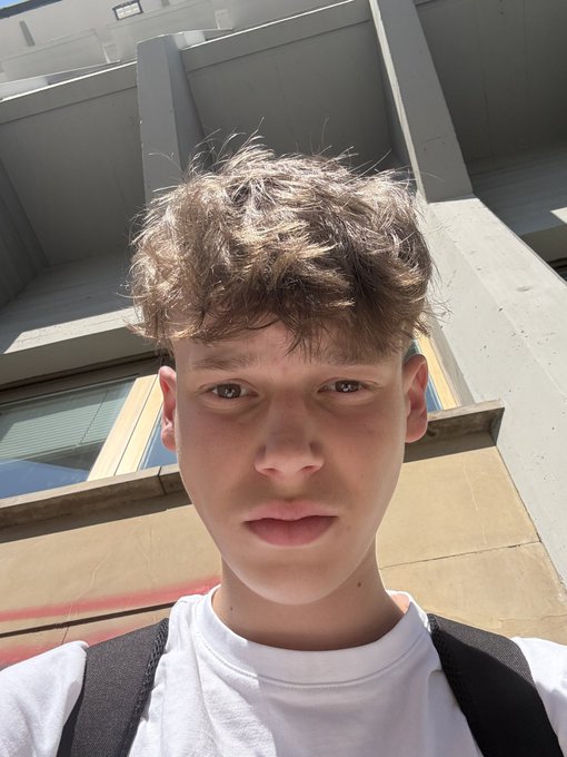 A selfie of a young man with curly brown hair wearing a white shirt, standing outside near a building with a concrete structure and windows in the background.