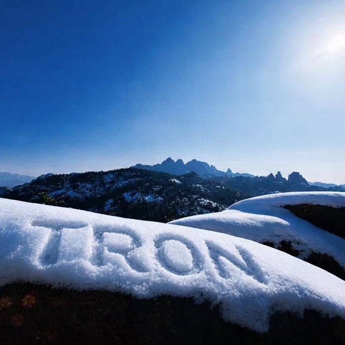 A snowy mountain landscape with the word 'TRON' written into the snow in the foreground, with rocky mountain peaks visible in the distance under a clear blue sky.
