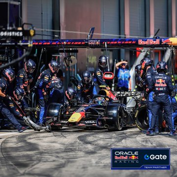 A Red Bull Racing pit crew in dark blue uniforms services a Red Bull Racing Formula 1 car in the pit lane, with team partner logos including "ORACLE Red Bull RACING," "Gate," and "Red Bull" visible, capturing the high-intensity teamwork at the Austrian Grand Prix weekend that the post highlights.