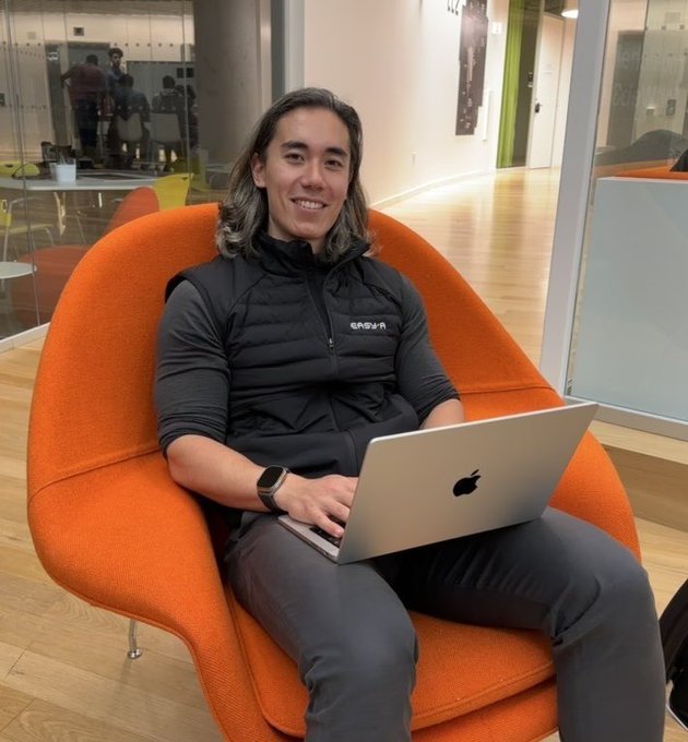 A smiling young man with long hair sits in a bright orange chair in a modern office lounge, working on a silver MacBook while wearing a black vest labeled 'Cognira' over a long-sleeve shirt.