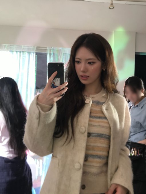 A young woman with long dark hair takes a mirror selfie with her smartphone in a room with white curtains, wearing a striped shirt and a fuzzy cream-colored cardigan.
