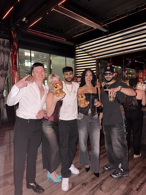 A group of five people posing together at what appears to be a rooftop bar, with two holding teddy bear-shaped drinks.