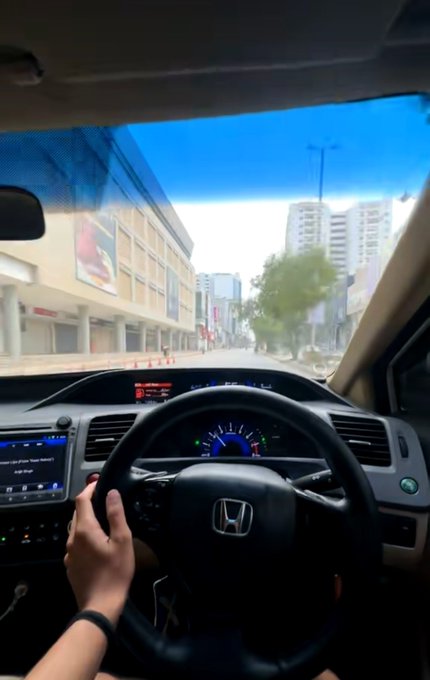 A first-person view from inside a Honda vehicle, showing the steering wheel, dashboard, and a city street view through the windshield.