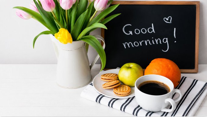 A cheerful "Good morning!" is written on a chalkboard beside a breakfast spread of tulips, biscuits, an apple, an orange, and a cup of coffee—setting a warm, motivational tone that matches the post's greeting and its upbeat call to continue the day's hustle for the family.