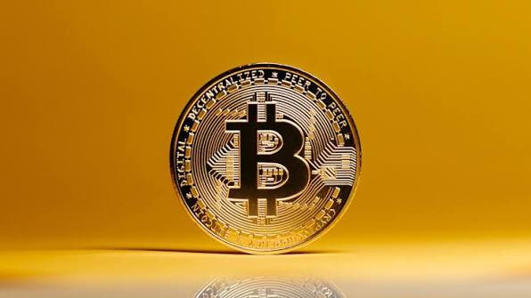 A close-up of a gold-colored physical Bitcoin token standing upright on a reflective surface against a yellow background, featuring the iconic 'B' symbol.
