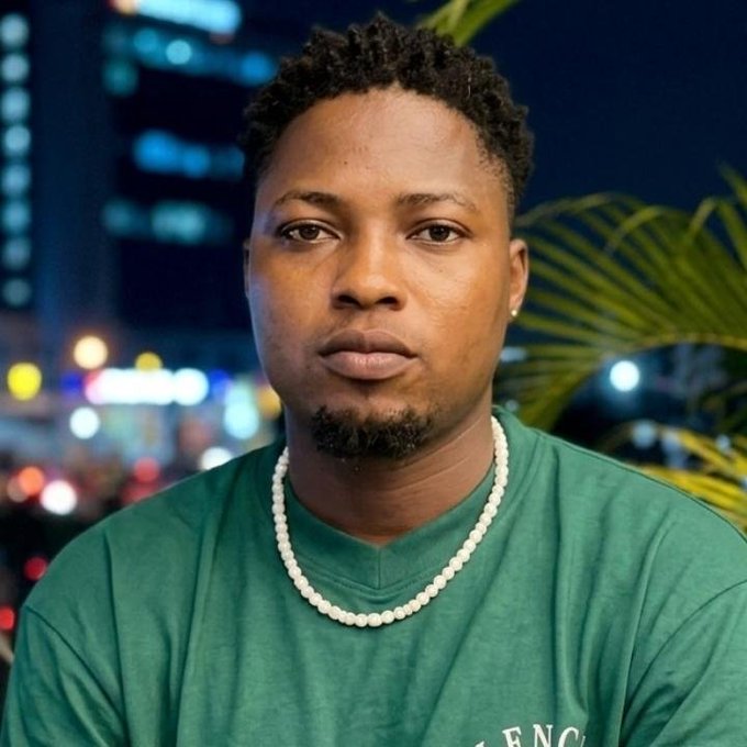 A portrait of a man with short curly hair and a goatee, wearing a green t-shirt and a pearl necklace, photographed at night with city lights in the background.