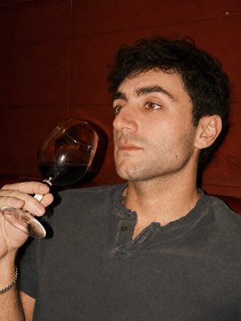 The image shows a dark-haired man wearing a gray henley shirt, holding a glass of red wine up toward his face as if inspecting or about to take a sip, with his eyes looking directly at the camera against a dark red background. The image humorously relates to the accompanying post about a cryptocurrency trader whose ultimate buy signal is seeing the "qwerty" (a slang term for a flaccid penis) appear as a size reference in a coin's chart—tying into the suggestive nature of the visual and the joke about sizing.