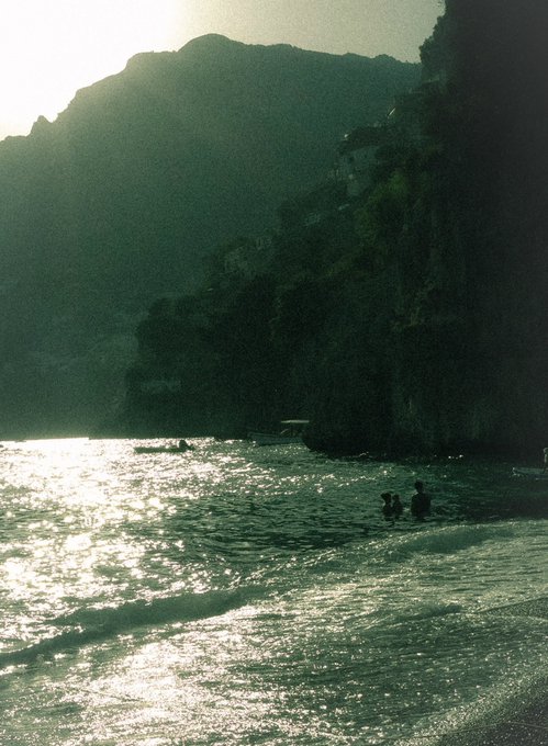Silhouetted figures wade in the glistening waters of a cove framed by steep, shadowed cliffs, capturing a serene twilight moment along the Amalfi Coast. The hazy, emerald-toned atmosphere and shimmering sea evoke the tranquil "goodnight" mood suggested in the post.