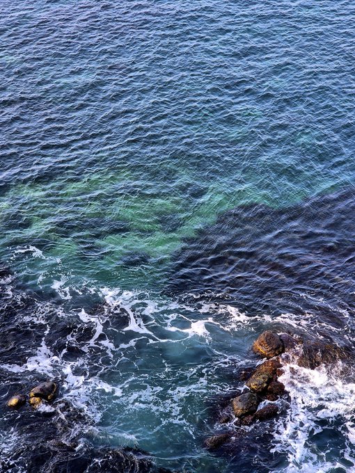 Aerial view of clear blue-green ocean water with white waves crashing against dark rocks along a rocky shoreline.