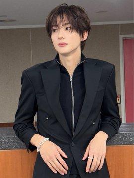 A young man with dark, tousled hair poses in a stylish all-black ensemble, wearing a tailored blazer over a high-neck zipped top. He accessorizes with a silver bracelet and a ring, posing against a neutral indoor background.