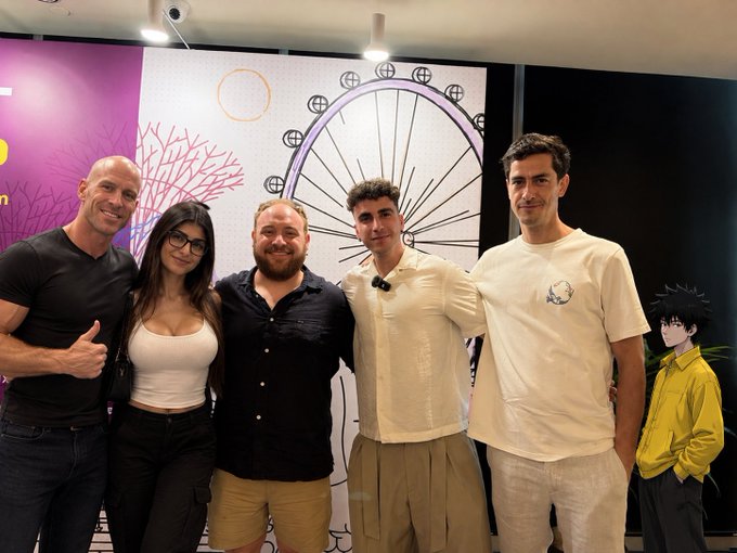 A group photo of five people posing together at what appears to be a Ghibli-themed event, standing in front of a wall featuring an illustration resembling a Ferris wheel and Totoro-style design, with a small anime figure on the right.