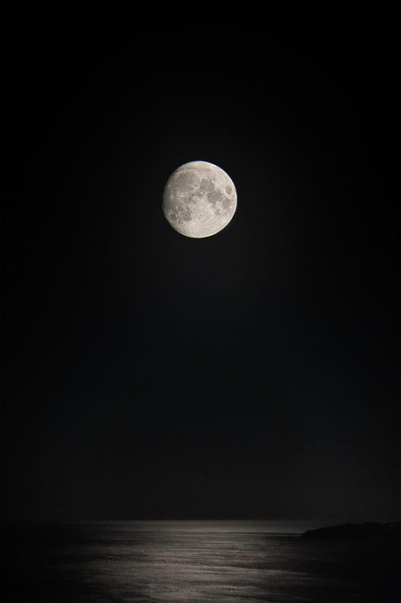 A bright full moon glowing in a dark night sky, with its light reflecting across a calm ocean below.