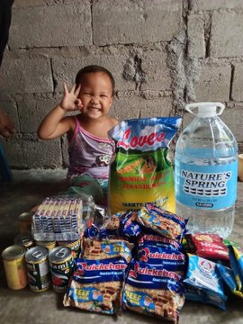 A smiling young girl makes an "OK" hand gesture beside a stack of donated supplies—including "Natures Spring" water jugs, "Skyflakes" crackers, "Boy Bawang" nuts, canned goods, and a bag of rice—reflecting the MEXC Foundation and Mochi Web3's relief efforts that provided food and clean drinking water to around 300 displaced families following the Mindanao earthquake.
