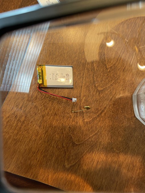 A photograph of a small lithium polymer battery with red and black wires attached to a JST connector, lying on a wooden surface.