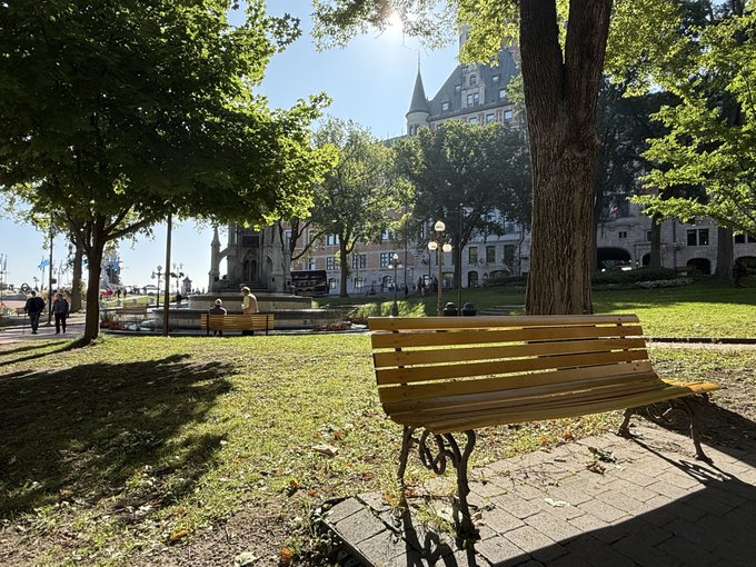 A sunlit park scene features a yellow wooden bench on a brick pathway, with large green trees, a historic building in the background, and a fountain area visible on the left.