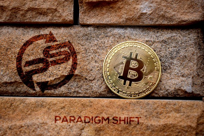 A graphic showing a transition from fiat to digital currency, with a recycling-style dollar sign next to a Bitcoin coin on a brick surface, captioned 'PARADIGM SHIFT'.