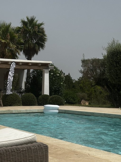 A white inflatable float drifts in a sun-drenched pool surrounded by palm trees and Mediterranean greenery in Ibiza, capturing the leisurely "sideways summer" the poster describes while waiting for the next bull market to accumulate BTC.