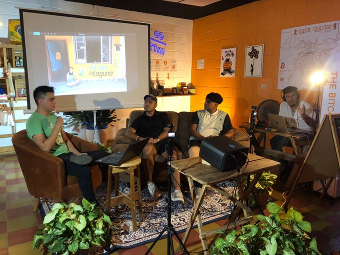 Four men sit in a podcast or panel setup in a room with Bitcoin/crypto-themed decorations, with a screen displaying 'bilaguna' and a map titled 'THE BITCOIN' visible on the wall.