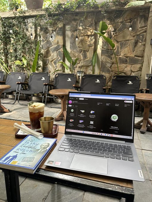 The image shows a laptop computer sitting open on top of a wooden table, with a book next to it. The laptop screen displays a website called "www.axisrobotics.com" along with a message that reads "GAxis (✱,✱) New week. New challenges. New access. We're giving away 10 BitRobot access codes over the next 72 hours. Winners will get access to SN/04 and start earning rewards from both Axis and BitRobot." The laptop is placed on a table surrounded by chairs, suggesting that it might be in an outdoor setting like a cafe or a park.