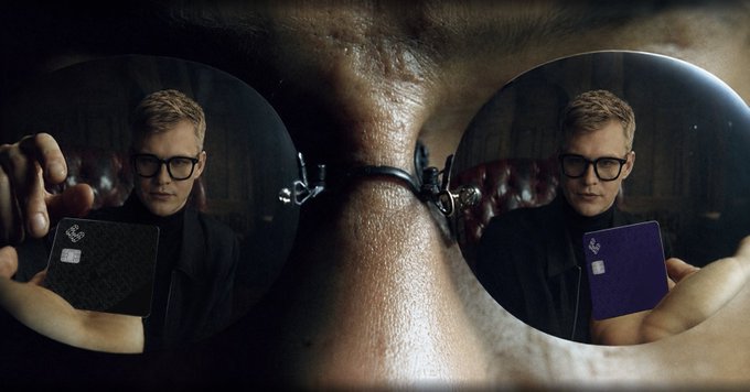 A close-up of a person wearing sunglasses, with the reflection of a man holding a card visible in both lenses.