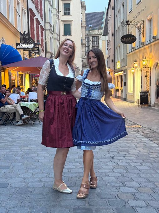 Two women wearing traditional Bavarian dirndls—one in a black top with red skirt, the other in a blue dress—pose smiling on a cobblestone street with outdoor cafes and a "GILARDI" sign visible in the background.