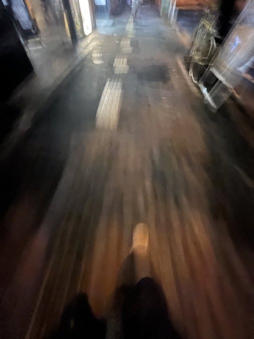 The image shows a person's feet walking down a street at night, with their shadow visible on the ground. The photo appears to be taken from behind them, giving an impression of motion blur as they walk away from the camera. The background features various objects such as a shopping cart and a suitcase, suggesting that this scene takes place in a busy urban area or market.