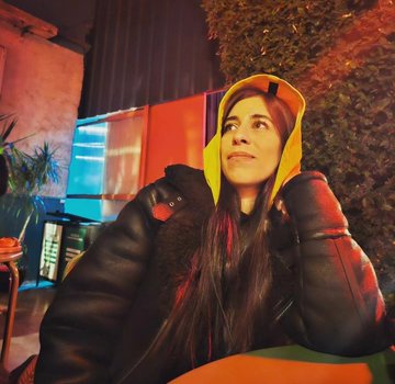 A woman with long dark hair wearing a yellow hooded jacket over a dark puffer coat looks up at night in a warmly lit outdoor setting.