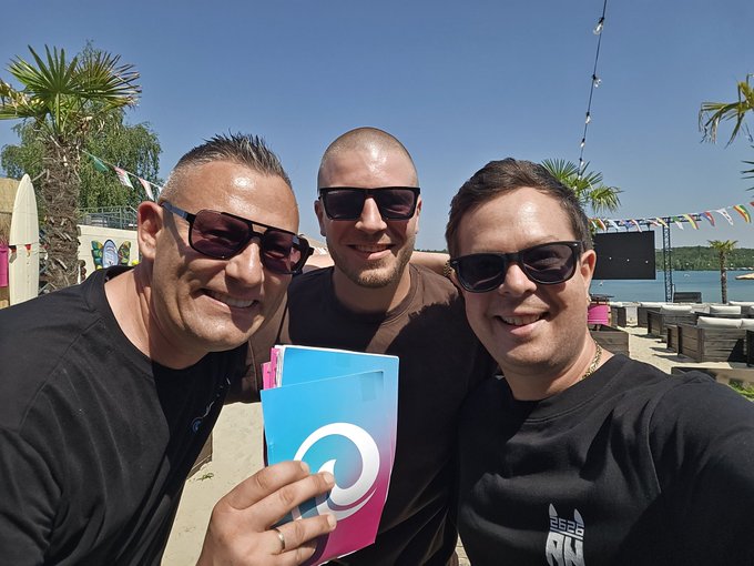The image shows three men posing for a photo, with one of them holding up a blue book that has a white logo on it. The background features palm trees and a beach setting, suggesting they are at an outdoor event or gathering. The caption "Crypcon Content continues A bit XRP-flavored this time" implies that the content being shared might be related to cryptocurrency or digital currency, possibly as part of a promotional campaign for a new cryptocurrency or a discussion about its potential uses and implications.