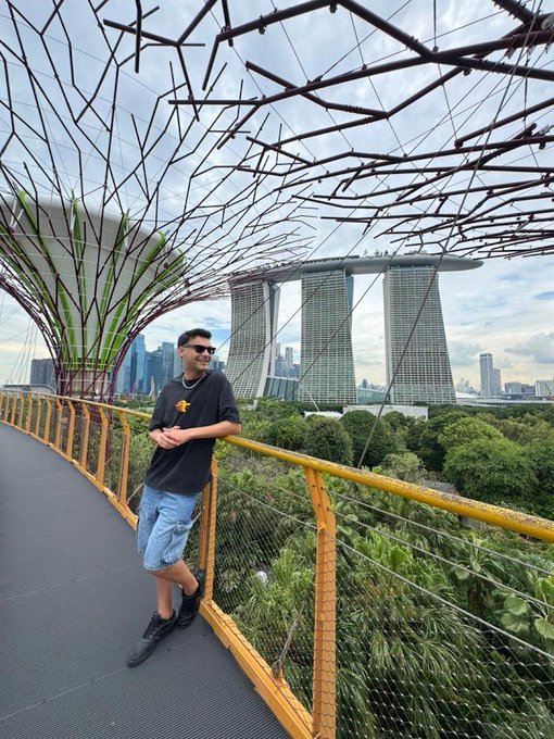 A photo of a man wearing sunglasses, a black t-shirt, and blue jeans, leaning against a yellow railing on an elevated walkway with a trellis structure, with the Marina Bay Sands hotel visible in the Singapore skyline behind him.