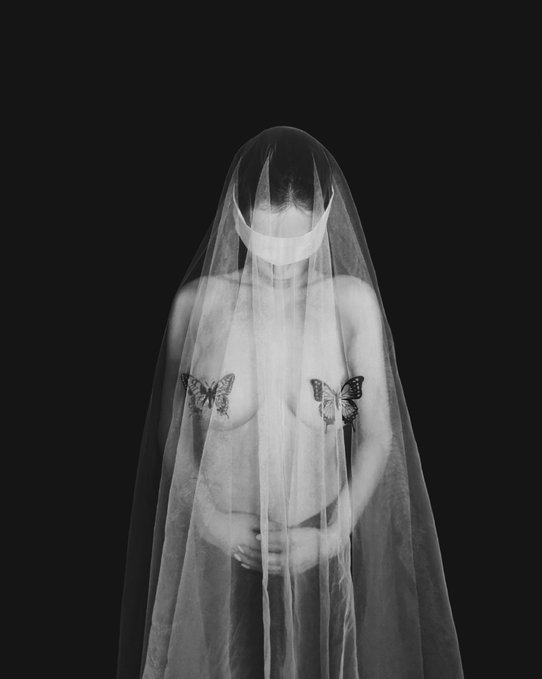 A black-and-white self-portrait of a topless figure draped in a sheer veil that obscures their face, with butterfly tattoos on both breasts and hands clasped at the waist, evoking the post's theme of hidden vulnerability and quiet mourning.