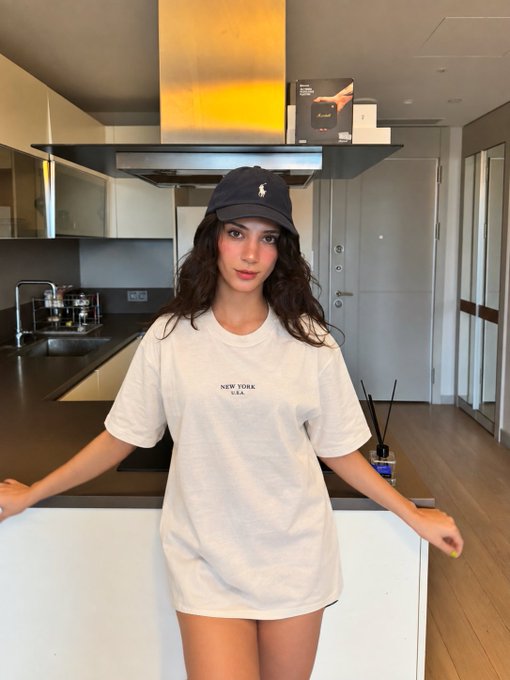 A young woman wearing a navy blue baseball cap and an oversized white t-shirt with 'NEW YORK U.S.A.' printed on it stands in a modern kitchen.