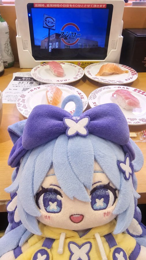 A plush doll with light blue hair and a purple bow sits in front of plates of sushi on a Kura Sushi (くら寿司) conveyor belt, with the restaurant's logo displayed on a screen above.