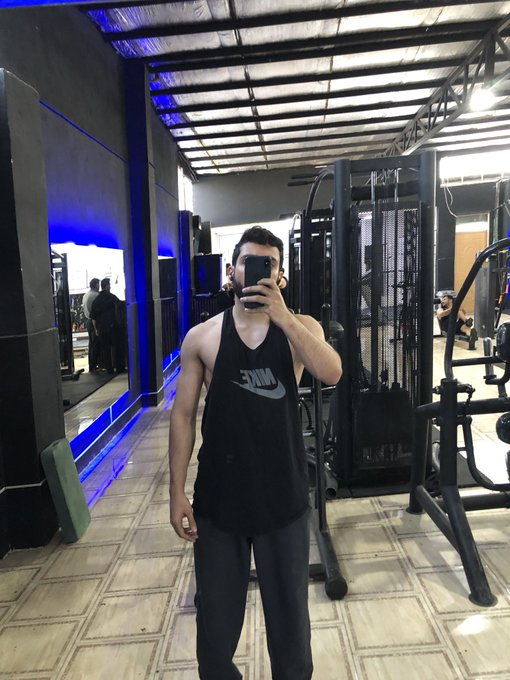 A mirror selfie of a fit young man in a black Nike tank top at a gym with equipment visible in the background.