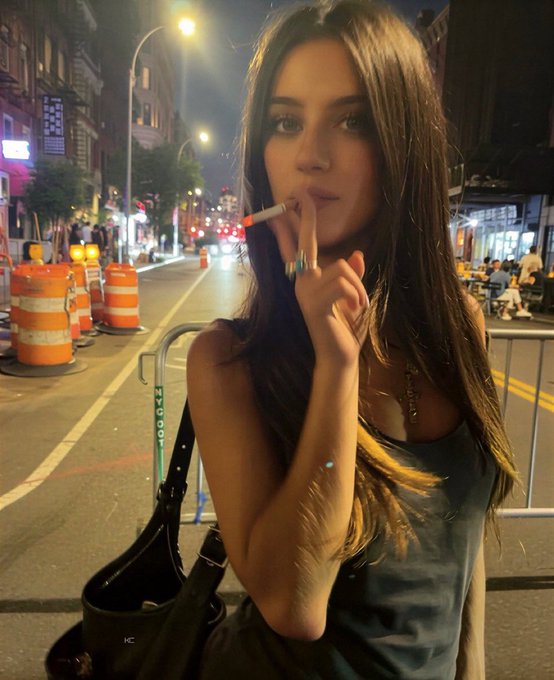 A young woman with long brown hair stands on a city street at night, smoking a cigarette, wearing a gray tank top and carrying a black bag, with orange construction barrels and a barricade visible behind her.