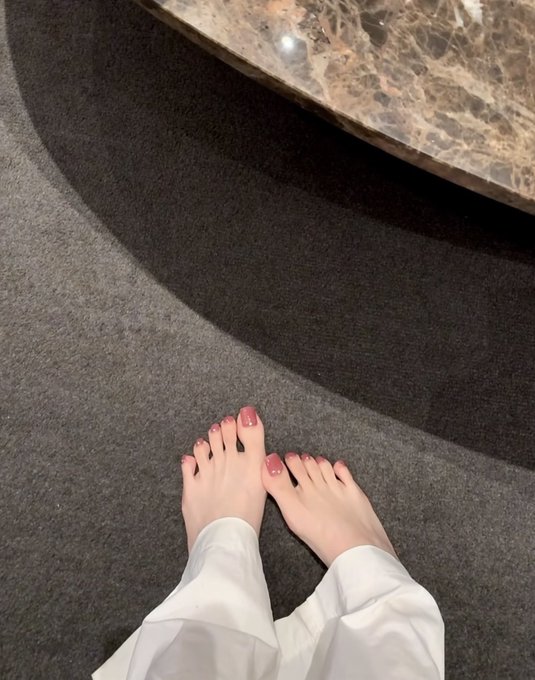 A downward view shows a person's bare feet with pink toenail polish standing on a dark gray carpet next to a marble surface.