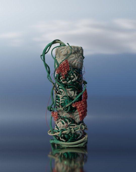 A cylindrical sculpture wrapped in textured fabric and tangled red and green threads stands against a soft blue and gray background with a reflective surface below.