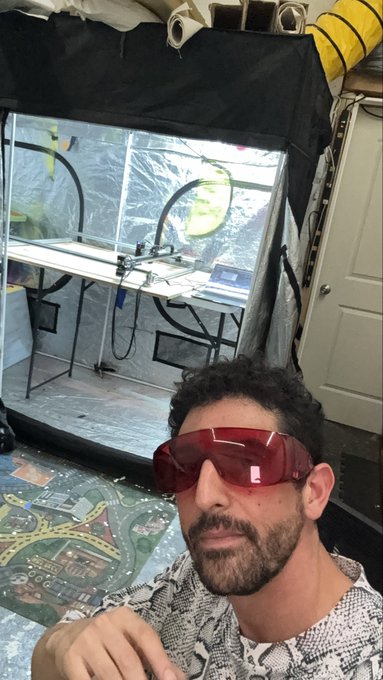 A bearded man wearing red safety goggles poses for a selfie inside a workshop containing a CNC machine and other equipment.