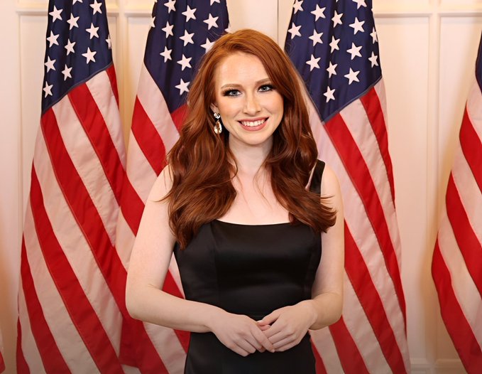 A woman with long red hair in a black dress stands smiling in front of several American flags.