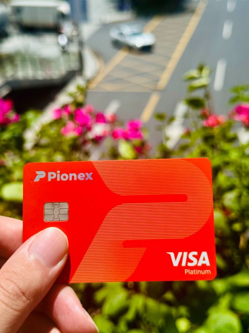 A hand proudly holds up a red Pionex Visa Platinum card with its visible chip, set against a blurred backdrop of a sunny street and blooming pink flowers—visually reinforcing the message that "the future is in our hands" by placing modern financial technology literally in the palm of a hand.