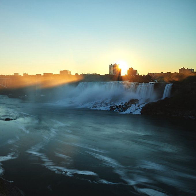 A serene sunset view of Niagara Falls, with the sun rising behind a distant city skyline and mist rising from the cascading water.