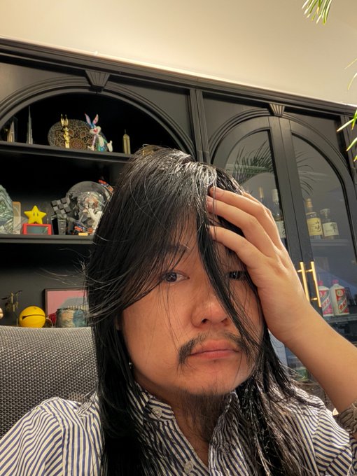 The image shows a person with long dark hair and a mustache, wearing a striped shirt, with their hand resting on their forehead in an exasperated or stressed expression, set against a backdrop of shelves displaying various collectibles and figurines. This visual conveys frustration or weariness, aligning with the post's message about stepping back from giving coin trading advice to avoid the stress and responsibility that comes with it.