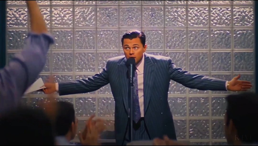 Leonardo DiCaprio, playing Jordan Belfort in 'The Wolf of Wall Street', stands at a microphone in a blue pinstripe suit with arms outstretched in front of a glass block wall.