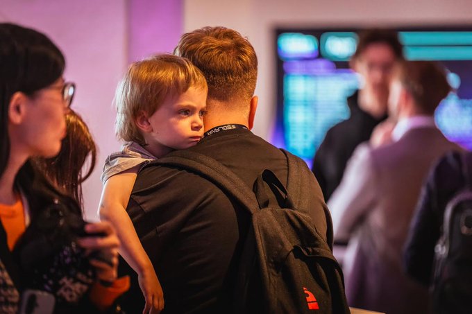 The image shows a man holding a small child, who appears to be tired from walking around at an event. The man has a backpack on his back, which suggests that they have been attending an event or gathering for some time. In the background, there are other people present as well. A large screen is visible in the scene, possibly displaying information about the event or related topics.