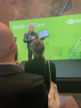 A photograph taken from the audience at an NVIDIA event, showing a man with white hair presenting on stage with a laptop in front of a large green screen displaying "Build-a-Claw."